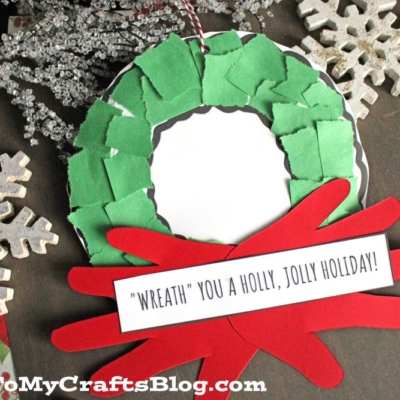 A handmade torn paper Christmas wreath craft made from green paper pieces, a red bow, and a white center with a note reading “‘Wreath’ you a holly, jolly holiday!”