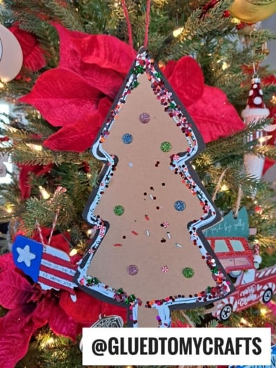 A handmade Paper Gingerbread Tree Craft ornament, decorated with glitter, sequins, and painted details, hangs on a Christmas tree with red poinsettias in the background. @GLUEDTOMYCRAFTS is written at the bottom.