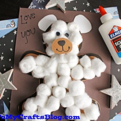 A cotton ball polar bear made of paper is glued to a dark background with handwritten text that says Love you beary much. A bottle of Elmer's glue and decorative stars are also visible.