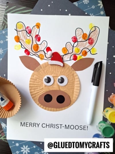 A cupcake liner Christmas moose craft made from a cupcake liner, colored antlers with fingerprint lights, paper ears, and a mini Santa hat, with MERRY CHRIST-MOOSE! written below. Art supplies surround this adorable cupcake liner moose creation.