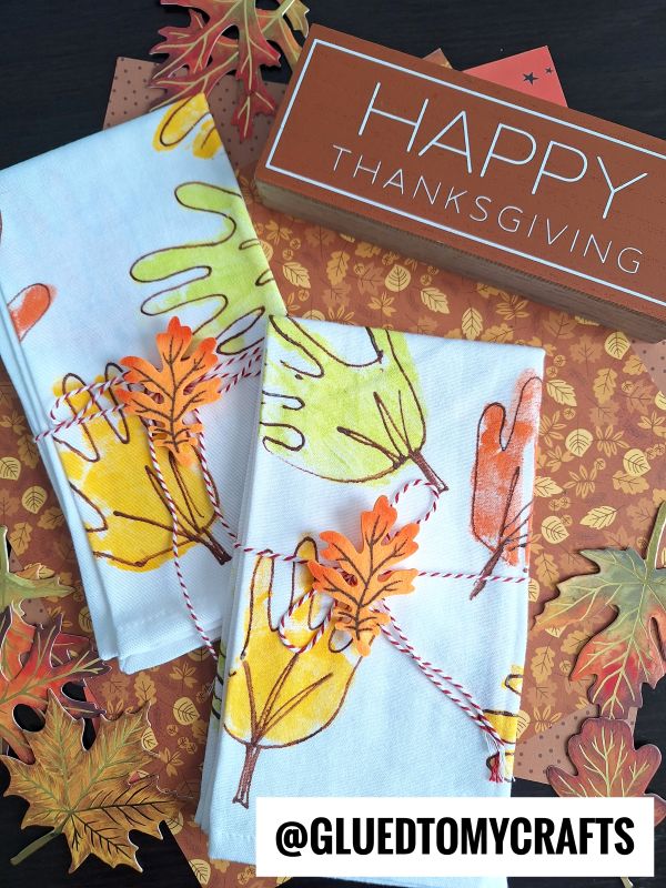 Hand towels decorated with colorful autumn leaves and Thanksgiving handprint crafts are displayed on a fall-themed surface with paper leaves. A sign reading HAPPY THANKSGIVING is in the background.