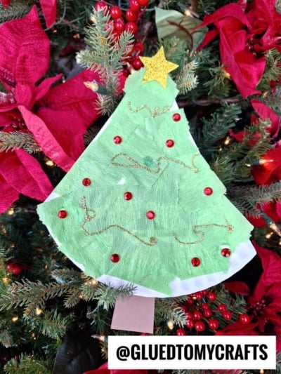 A Paper Plate Christmas Tree craft with green paint, red sequins, and gold glitter glue, topped with a yellow star, is displayed on a Christmas tree with red poinsettias. @GLUEDTOMYCRAFTS is written at the bottom.