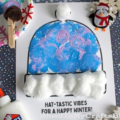 A children’s winter craft shows a pom pom stamped winter hat painted blue and purple, decorated with cotton balls on a printed page reading “HAT-TASTIC VIBES FOR A HAPPY WINTER!” Surrounded by art supplies, snowflakes, and a small penguin plush.