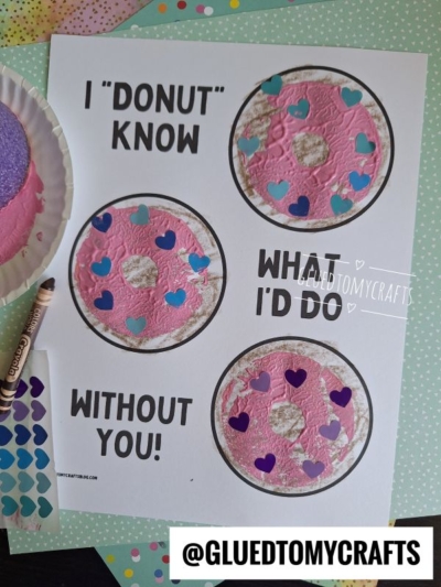 A craft sheet with three pool noodle stamped donuts decorated with pink paint and heart stickers, alongside the text I DONUT KNOW WHAT ID DO WITHOUT YOU! Crayons and crafting materials are nearby.