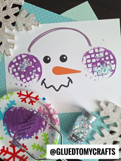 A snowman face craft on white paper, featuring a drawn orange carrot nose and purple potato masher stamped ear muffs made with paint and glitter. Craft materials and snowflake decorations surround the artwork.