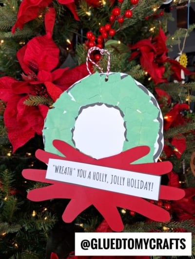 A handmade torn paper Christmas wreath craft using green and red paper hangs on a tree with red poinsettias. A label reads “‘Wreath’ you a holly, jolly holiday!” and @GLUEDTOMYCRAFTS is displayed at the bottom.