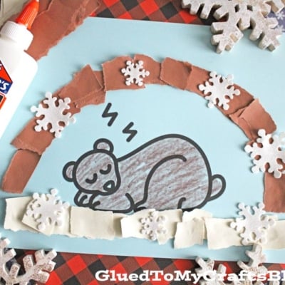 A craft features a torn paper hibernating bear sleeping under a cozy torn paper bear cave made of brown and white strips, accented with snowflakes. A glue bottle and patterned paper sit in the background.