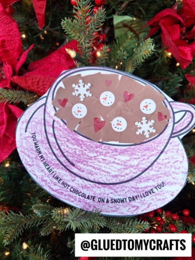 A torn paper hot chocolate mug filled with snowman and snowflake marshmallows sits on a Christmas tree. The cup reads: You warm my heart like hot chocolate on a snowy day! I love you. @GLUEDTOMYCRAFTS.