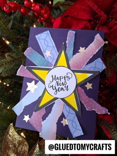 A festive handmade New Year card with a central yellow star, layered with torn paper firework strips and small gold stars. "Happy New Year" is written in the center. The card is displayed on a Christmas tree.