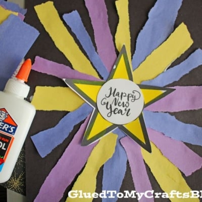A New Year’s craft with a torn paper firework made of yellow and purple rays on black paper, a star in the center reading “Happy New Year,” and a bottle of Elmer’s glue