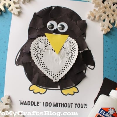 A torn paper penguin craft made with black paper, a doily heart, googly eyes, and a yellow beak sits on a sheet that says WADDLE I DO WITHOUT YOU?! Nearby are snowflake decorations and a bottle of glue.