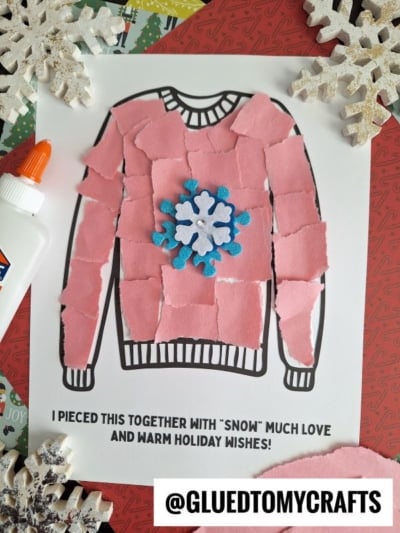 A torn paper winter sweater craft features a sweater shape on white paper, covered with pink torn paper pieces and a blue-and-white snowflake in the center. A glue bottle, snowflake decorations, and a heartfelt holiday message complete the scene.