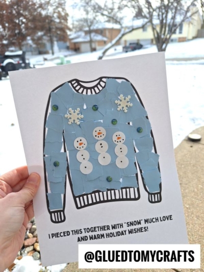 A hand holds a torn paper winter sweater craft, decorated with snowmen, snowflakes, and sequins, with text that reads, I pieced this together with snow much love and warm holiday wishes! Snowy yard in the background.
