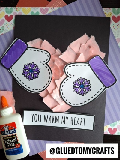 Two paper mittens with purple cuffs and snowflake designs rest over a torn paper heart on black paper. A glue bottle and cut-out text reading YOU WARM MY HEART complete this heartfelt scene.