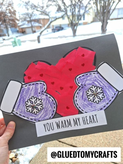 A hand holds a craft featuring a red torn paper heart with small hearts, two purple mittens with snowflake designs, and the phrase "You warm my heart" on black paper. Snowy houses and trees are in the background.
