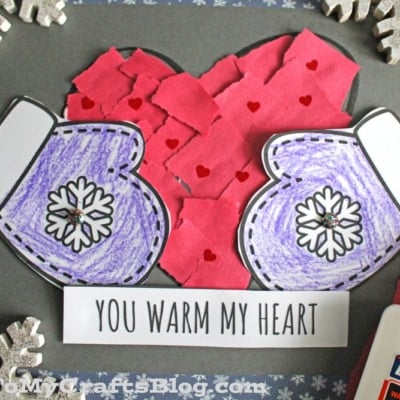 A handmade craft with purple mittens, a red heart crafted from torn paper, and the words You warm my heart. A glue bottle and snowflake decorations surround the craft.