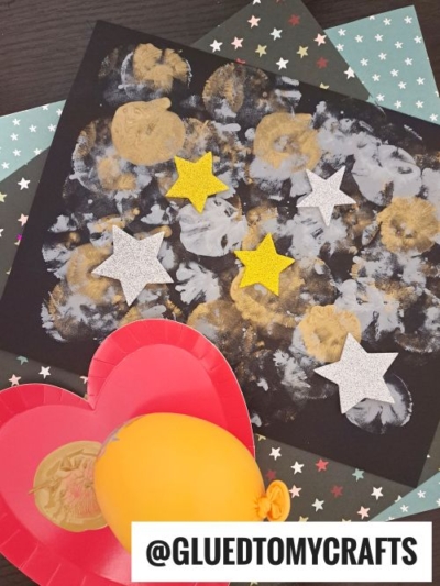 A black paper with gold and white painted circles, decorated with glittery white and yellow star cutouts—perfect for New Year’s Eve Crafts For Kids. Red heart-shaped plates, a yellow balloon, and star-patterned paper are nearby. @GLUEDTOMYCRAFTS at the bottom.