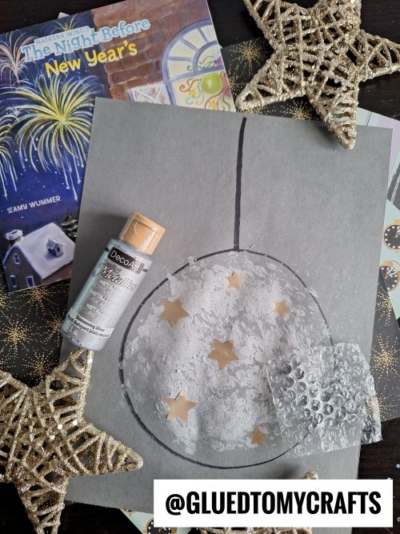A gray sheet displays a drawn ornament decorated with silver paint and star-shaped cutouts—perfect inspiration for New Year’s Eve Crafts For Kids—surrounded by gold stars, a bottle of silver paint, bubble wrap, and The Night Before New Year’s book.
