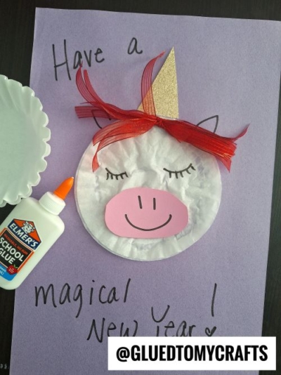 A unicorn face made from a white paper plate, pink paper snout, red and gold ribbon mane, and gold horn is glued to purple paper with Have a magical New Year! written on it—perfect for New Year’s Eve Crafts For Kids. Elmer’s glue is nearby.