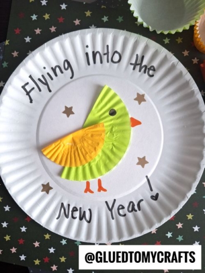 A fun New Year’s Eve Crafts For Kids idea: a paper plate craft featuring a yellow and orange cupcake liner bird, with drawn eyes, beak, and legs. “Flying into the New Year!” is written on the plate, decorated with star stickers and @GLUEDTOMYCRAFTS.