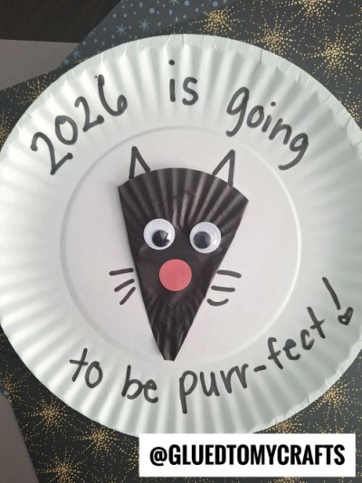 A paper plate craft shows 2026 is going to be purr-fect! written around a black cupcake liner cat face with googly eyes and a red nose. It’s one of the cutest New Year’s Eve Crafts For Kids! The @GLUETOMYCRAFTS credit is at the bottom.