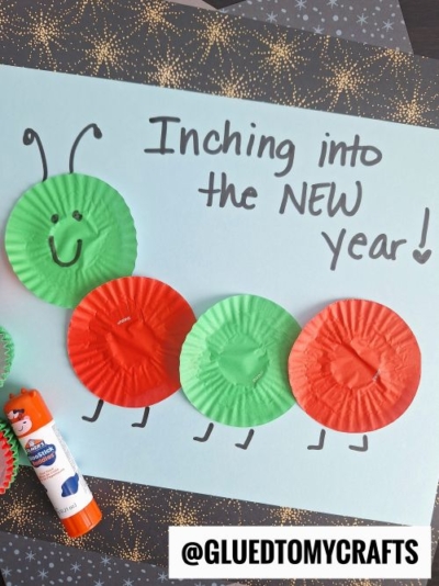 A craft of a caterpillar made from red and green cupcake liners on blue paper, with "Inching into the NEW year!" handwritten beside it. Perfect for New Year’s Eve Crafts For Kids! A glue stick and craft supplies are nearby. @GLUEDTOMYCRAFTS is at the bottom.