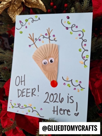 A handmade card features a reindeer face made from a tan cupcake liner, googly eyes, a red nose, and drawn antlers. It reads “OH DEER! 2026 is Here!”—a fun New Year’s Eve Crafts For Kids project with colorful designs.