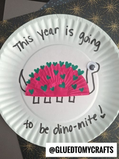 A fun New Year’s Eve Crafts For Kids project, this paper plate craft features a dinosaur made with a pink cupcake liner, green heart shapes, a googly eye, and the text “This year is going to be dino-mite!” around it.