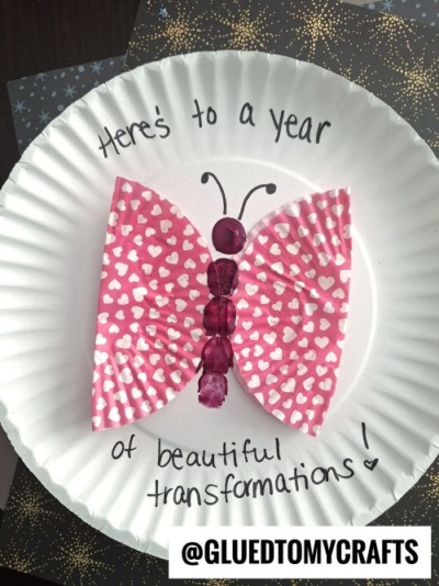 A paper plate displays a butterfly made from pink, heart-patterned wings, purple buttons as the body, and thumbprint accents. Above, handwritten text reads, Here's to a year of beautiful transformations! Perfect for New Year's Eve crafts.
