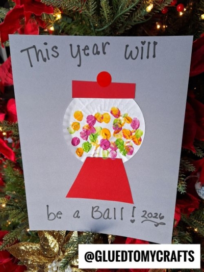 A handmade card resembling a gumball machine features thumbprint “gumballs” and the message “This year will be a ball! 2024,” making it a delightful New Year’s Eve craft displayed in front of a decorated Christmas tree.
