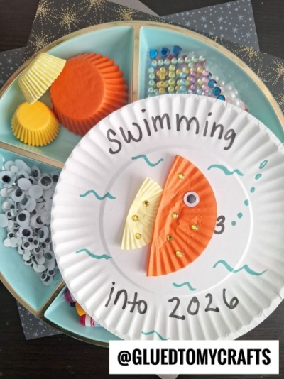 A paper plate craft features a fish made from orange and yellow cupcake liners, a googly eye, and gems—perfect for New Year’s Eve Crafts For Kids. Supplies include liners, beads, gems, and eyes. Text on the plate reads, Swimming into 2026.