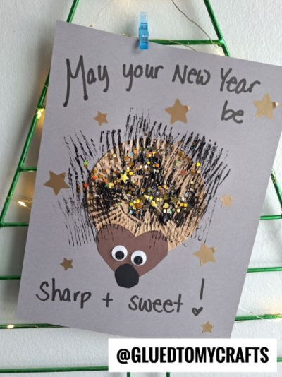 A handmade New Year card is a charming example of New Year’s Eve Crafts For Kids, featuring a hedgehog made from paint, glitter, googly eyes, and a brown paper nose. It reads "May your New Year be sharp + sweet!" and has gold star stickers.