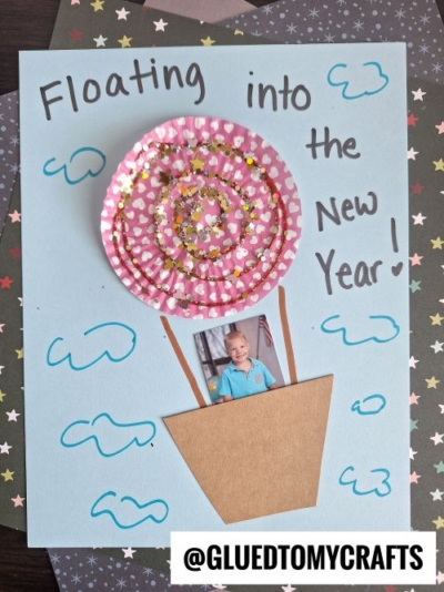 A child’s photo appears in a hot air balloon made from a paper cup liner with gold glitter, on blue paper with cloud doodles. Perfect for New Year’s Eve Crafts For Kids! The text reads: Floating into the New Year! @GLUEDTOMYCRAFTS tag at bottom.