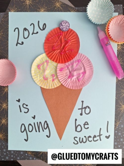 A paper craft of an ice cream cone made from colored cupcake liners and brown paper sits on light blue paper. Perfect for New Year’s Eve Crafts For Kids, “2026 is going to be sweet!” is written around it with a pink glitter pen and cupcake liners nearby.