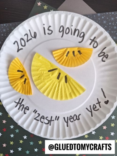 A paper plate decorated with three yellow citrus slices made from cupcake liners, perfect for New Year’s Eve Crafts For Kids, with "2026 is going to be the zest year yet!" written around them. The image is labeled @GLUEDTOMYCRAFTS.