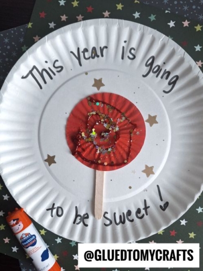 A fun New Year’s Eve Crafts For Kids idea: A paper plate decorated with star stickers and “This year is going to be sweet!” A red cupcake liner with glitter and a craft stick form a lollipop in the center. A glue stick sits nearby.