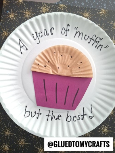 A paper plate craft featuring a construction paper muffin with a brown top and purple bottom—perfect for New Year’s Eve Crafts For Kids. It reads “A year of ‘muffin’ but the best!” above and below the muffin. @GLUEDTOMYCRAFTS is at the bottom.