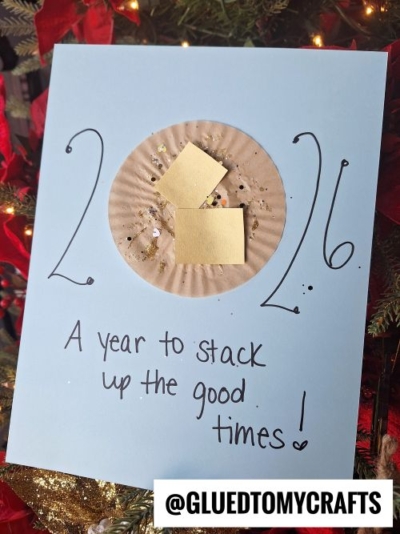 A blue card with “2024” written on it, featuring a brown cupcake liner decorated with gold paper squares and the words “A year to stack up the good times!”—a fun New Year’s Eve Crafts For Kids idea amid festive Christmas decorations.