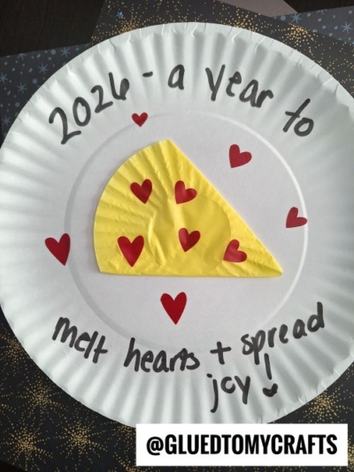 A paper plate with a yellow paper pizza slice, red hearts, and the handwritten message 2026 - a year to melt hearts + spread joy! Perfect for New Year’s Eve Crafts For Kids. The Instagram handle @GLUEDTOMYCRAFTS appears at the bottom.