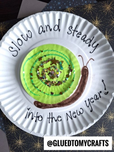 A paper plate decorated with a green spiral snail, glitter, and the words Slow and steady into the New Year! makes a fun New Year’s Eve Craft For Kids. A brown snail head is drawn, and @GLUEDTOMYCRAFTS is at the bottom.