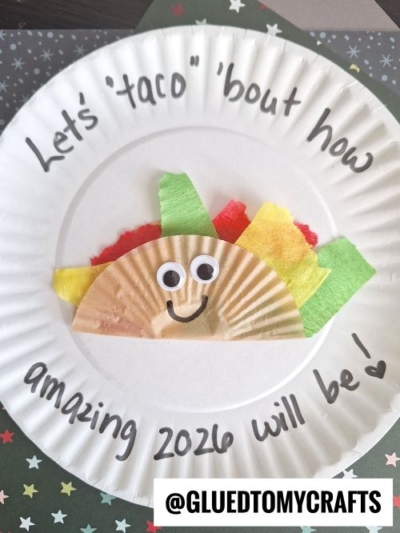 A paper plate with a smiling taco craft made of a cupcake liner and colored paper. Text around the plate reads, "Let’s taco ‘bout how amazing 2026 will be!" Perfect for New Year’s Eve Crafts For Kids. The handle @GLUEDTOMYCRAFTS appears at the bottom.