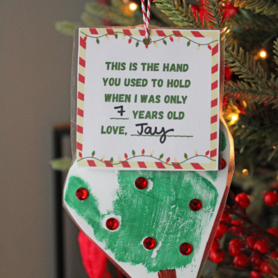 A handmade Christmas ornament features a child’s green handprint as a tree, red gems as ornaments, and a heartfelt card reading, “This is the hand you used to hold when I was only 7 years old. Love, Jay.”.