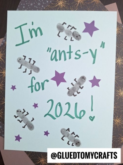 A handmade card reads Im ants-y for 2024! with purple stars and thumbprint ant drawings. The username @GLUEDTOMYCRAFTS appears at the bottom. Perfect for thumbprint New Year's Eve crafts, the card is placed on patterned paper.