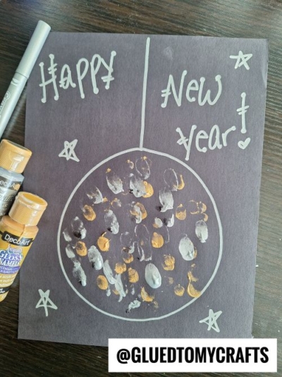 A handmade New Year card, perfect for New Year’s Eve Crafts For Kids, features "Happy New Year!" in silver on black paper, a large ornament with gold and white paint dots, hand-drawn stars, plus nearby paint bottles and a marker.