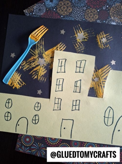 Yellow paper buildings with hand-drawn windows and doors are set before black paper decorated with yellow starburst fireworks, created using a plastic fork. This fun project is perfect for New Year’s Eve Crafts For Kids! The handle has @GLUEDTOMYCRAFTS printed on it.