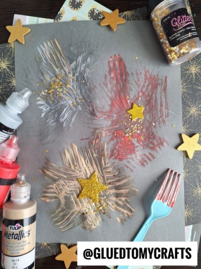 A gray paper with red, gold, and silver paint strokes made using a plastic fork, decorated with gold glitter and foam stars—it's a festive idea for New Year’s Eve Crafts For Kids. Paint bottles and glitter surround the sparkling artwork.