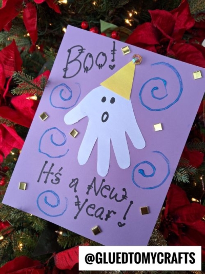 A festive New Year’s Eve craft for kids featuring a handprint ghost in a yellow party hat on purple paper, with "Boo! It's a New Year!" written in black, plus blue swirls and gold squares to decorate the page.
