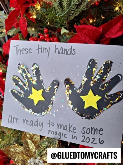 A festive New Year’s Eve craft features two black handprints decorated with gold stars and glitter on gray paper, with the text: “These tiny hands are ready to make some magic in 2026.” A Christmas tree is in the background.