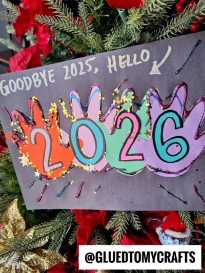 A colorful craft sign with handprint cutouts and the numbers 2026 makes a festive New Year’s Eve Crafts For Kids project. Above, text reads Goodbye 2025, Hello, with an arrow pointing to the numbers. Displayed on a Christmas tree. @GLUEDTOMYCRAFTS is at the bottom.