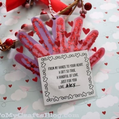 A handmade Valentine card shaped like a handprint with red and pink glitter, topped with heart stickers. A note in the center reads: “A handful of love… just for you. Love, Alex.”